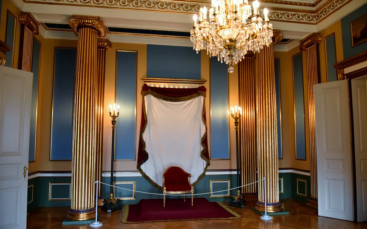 En sal på piano nobile på Amalienborg med riflede forgyldte søjler, en krystal lysekrone og en stol med baldakin på et rødt tæppe.