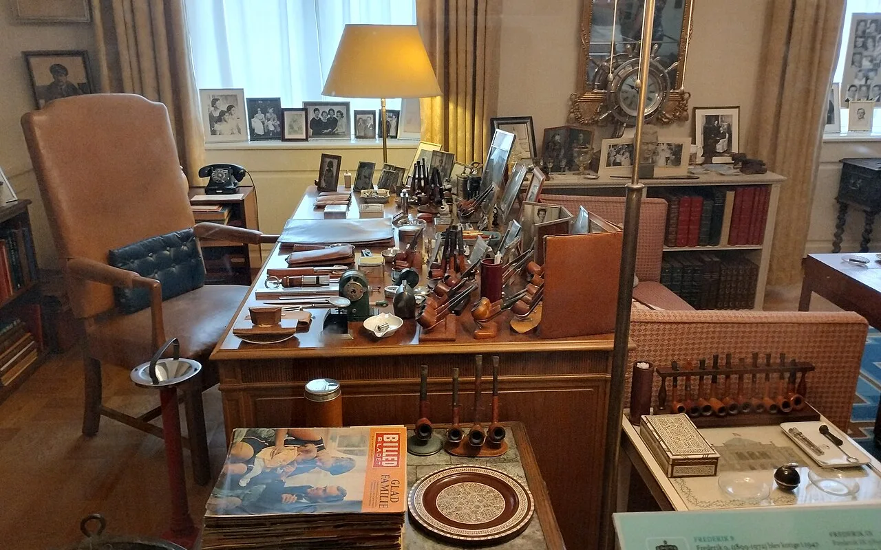 En konges skrivebord på Amalienborgmuseet, fyldt med piber, indrammede familiefotografier og to bordlamper.